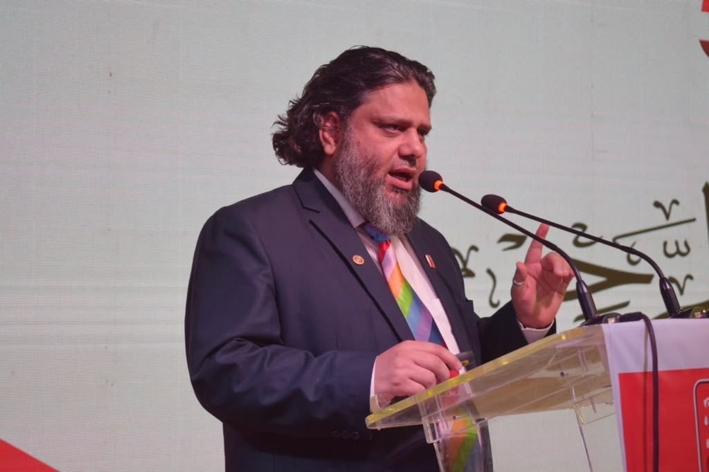 Dr. Ahmed Ejaz speaking at a podium in front of an Urdu calligraphy backdrop