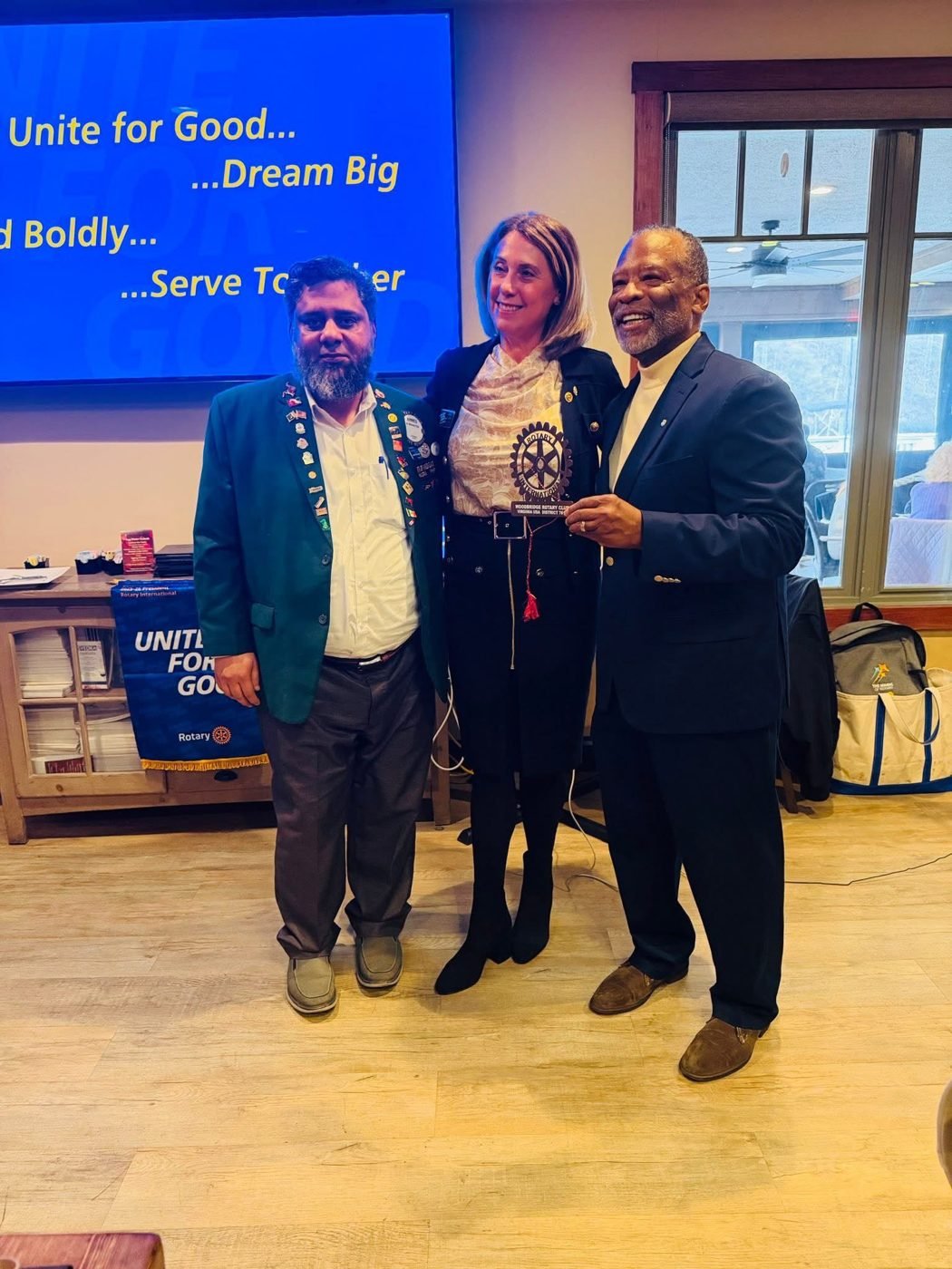 Dr. Ahmed Ejaz with two Rotarians holding a plaque under a Unite for Good, Dream Big, Serve Together screen
